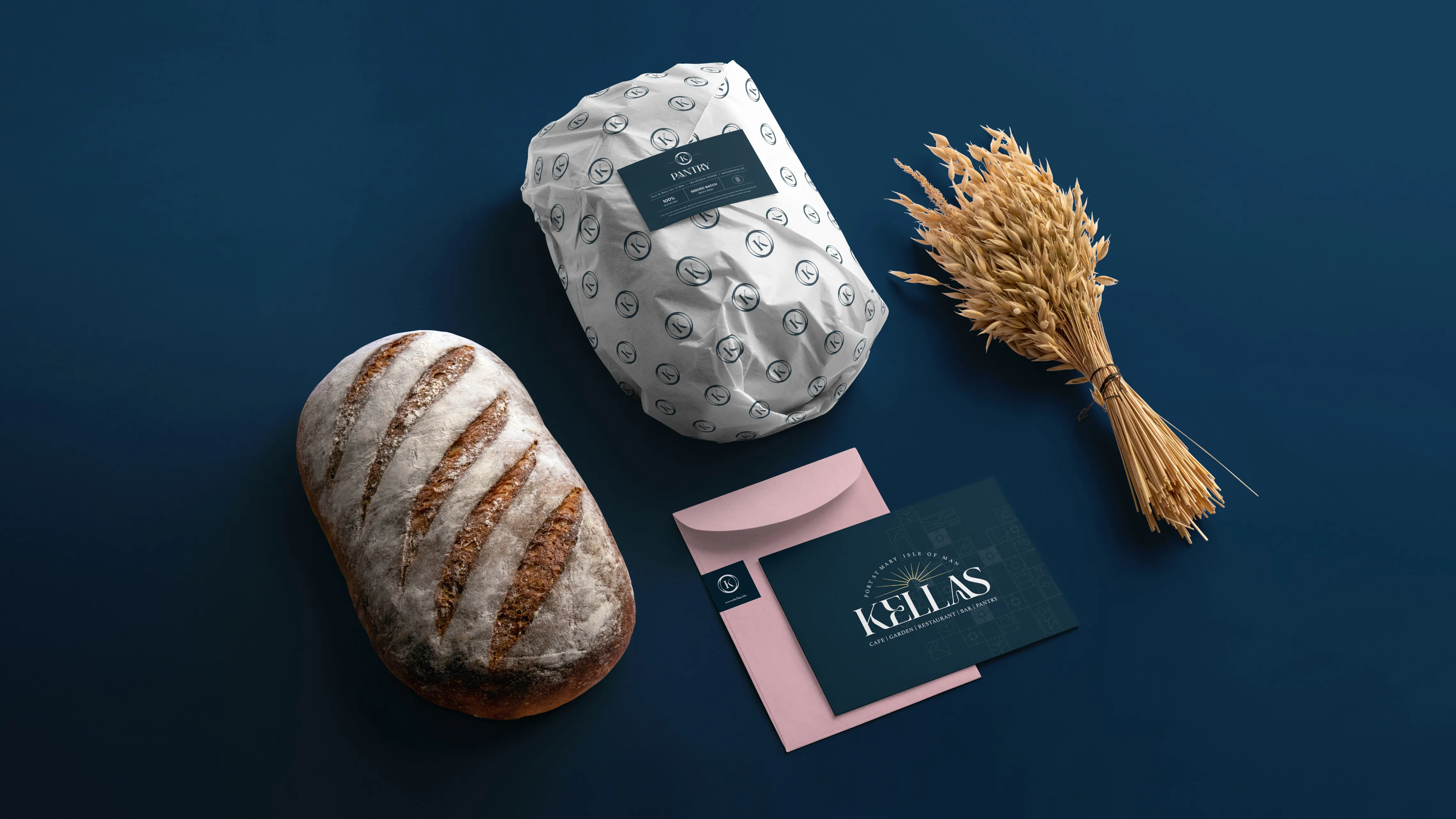 A styled flatlay on a dark teal background featuring a rustic sourdough loaf, a white branded pantry wrap/packaging with the Kella's pattern, a bundle of dried wheat, a pink envelope, and a dark teal Kella's business card.
